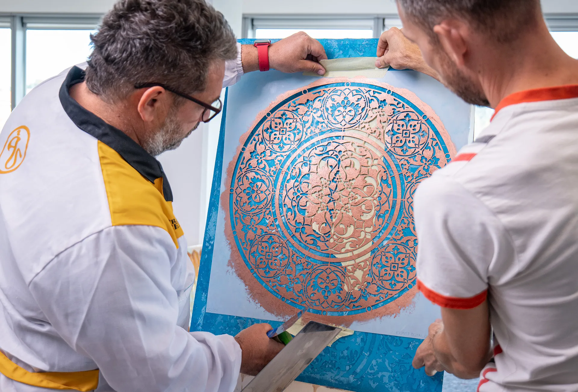 Due applicatori sollevano lo stencil da un decoro color rame su fondo azzurro